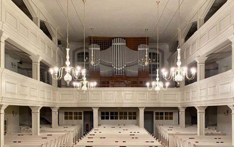 Weiße Innenansicht einer Kirche mit einer Orgel und mehreren Kronleuchtern.