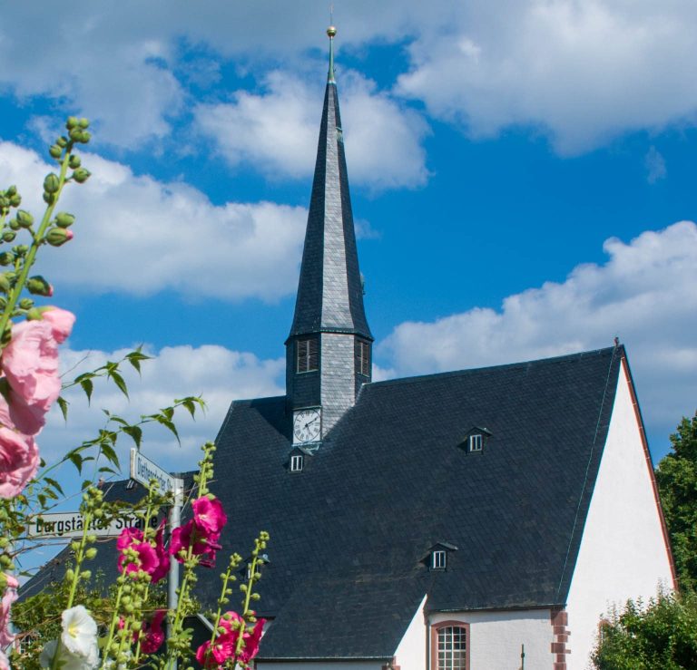 Kirche mit spitzem Turm, umgeben von bunten Blumen und blauen Himmel.