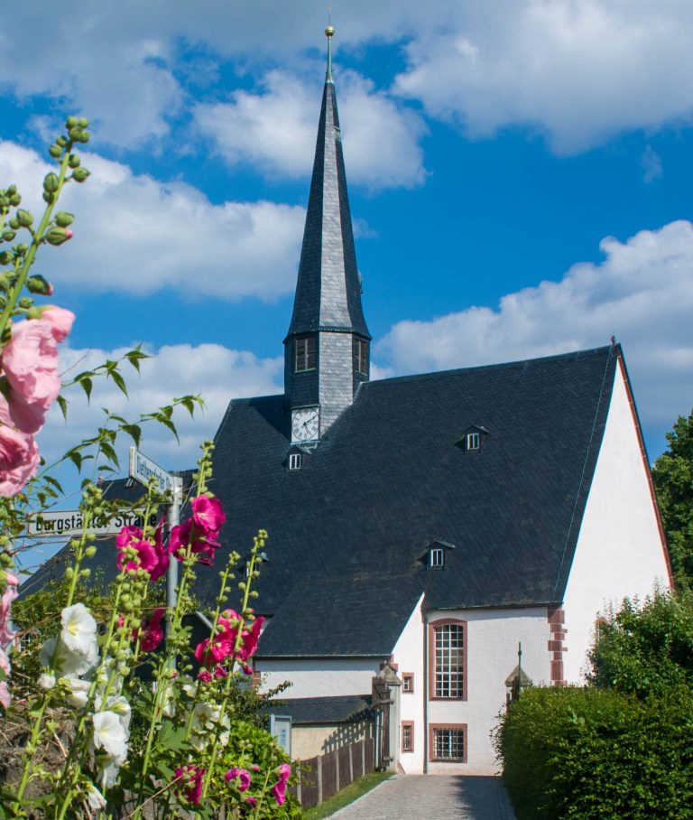 Kirche mit spitzem Turm, umgeben von Blumen und blauem Himmel.