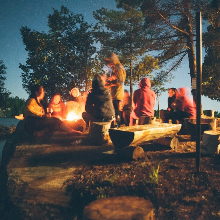 Eine Gruppe von Menschen sitzt um ein Lagerfeuer in der Natur bei Nacht.