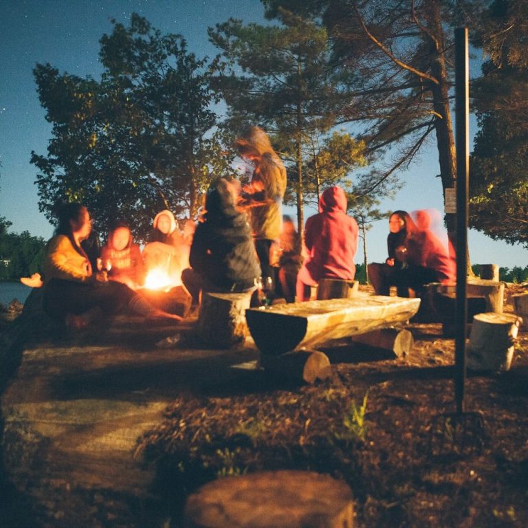 Gruppe von Menschen sitzt um ein Lagerfeuer in der Nacht, umgeben von Bäumen.