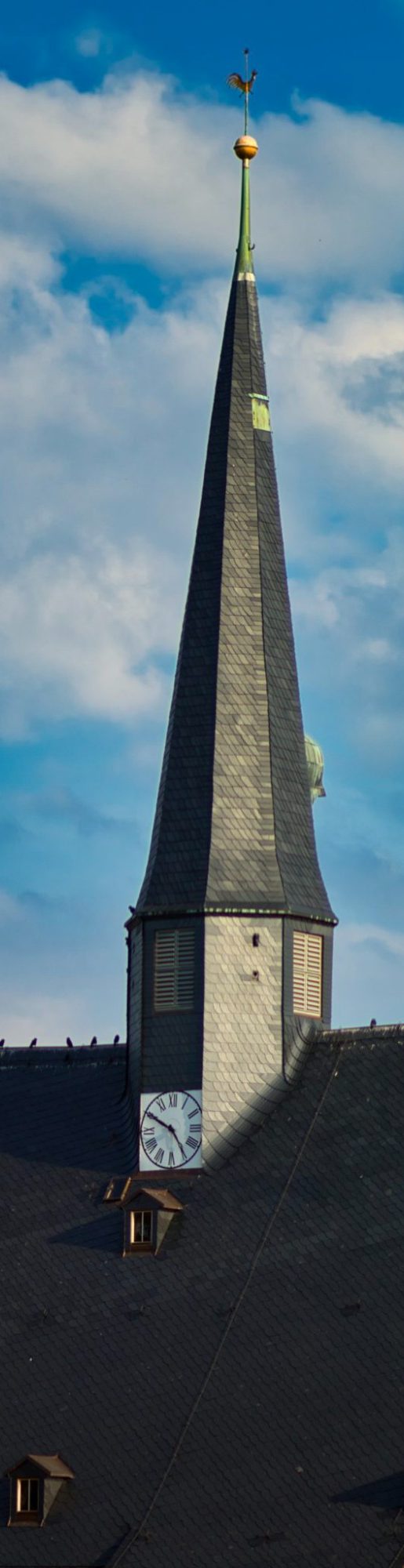 Spitzturm einer Kirche mit grauem Dach und blauem Himmel im Hintergrund.